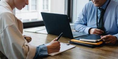 Image of 2 people in a meeting. One person is behind a computer and one is signing a document.